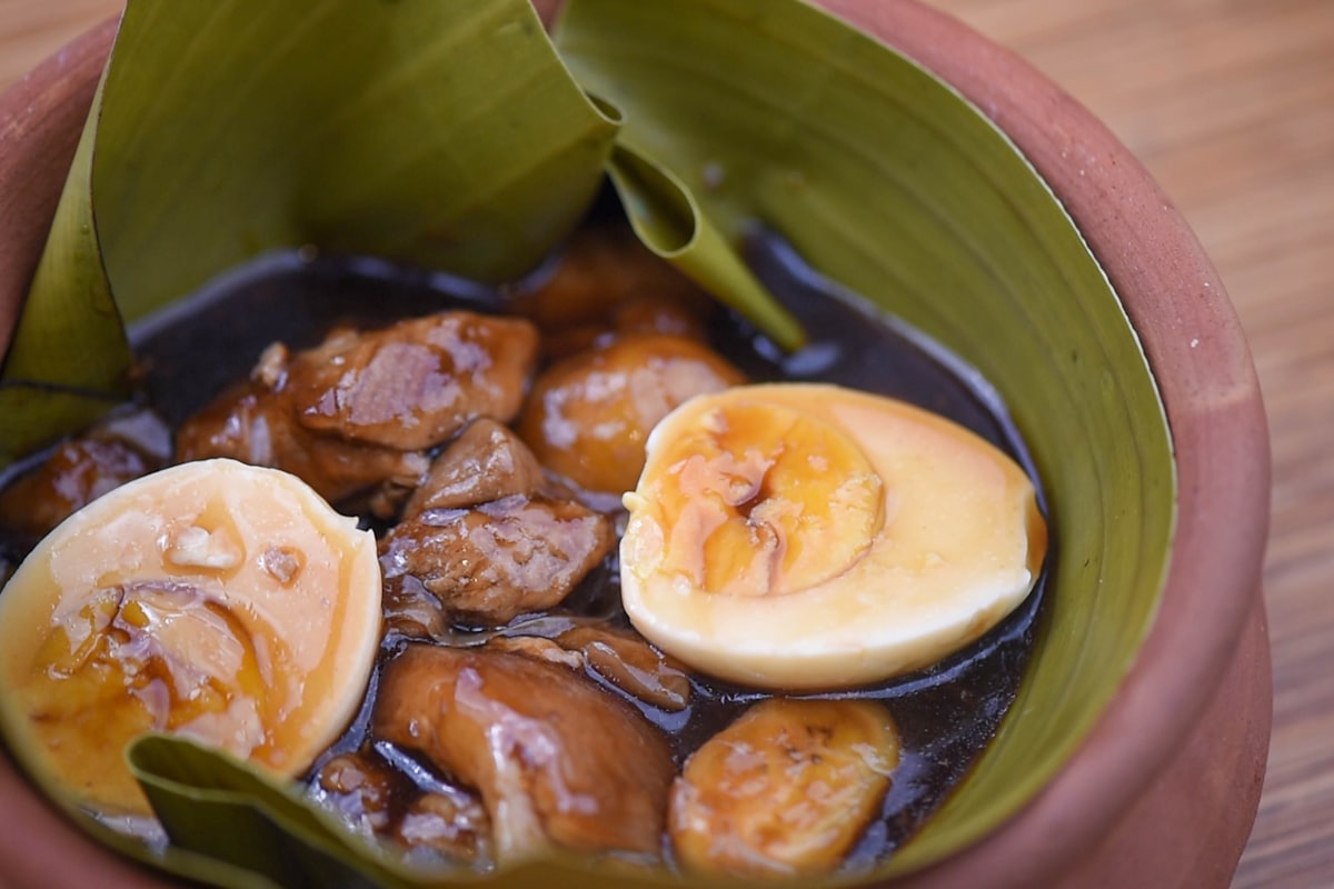 A pot of chicken adobo with dark glossy sauce, whole garlic cloves, bay leaves, and cracked peppercorns over steamed rice
