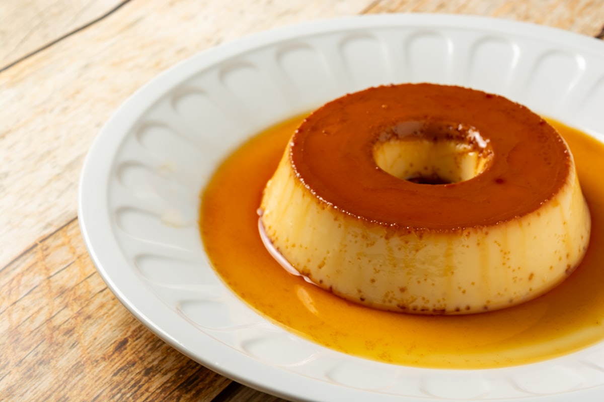 A slice of golden leche flan on a plate with dark caramel syrup pooling around the smooth custard